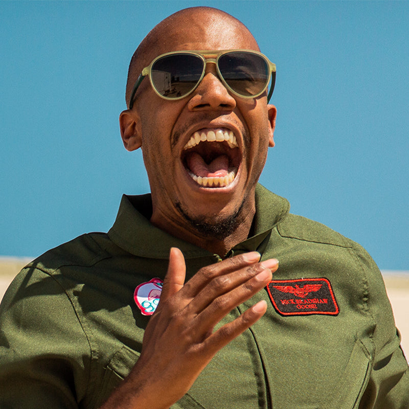 Un hombre con un mono verde de estilo militar y gafas de sol de aviador verde cadete con cristales verdes degradados grita alegremente.