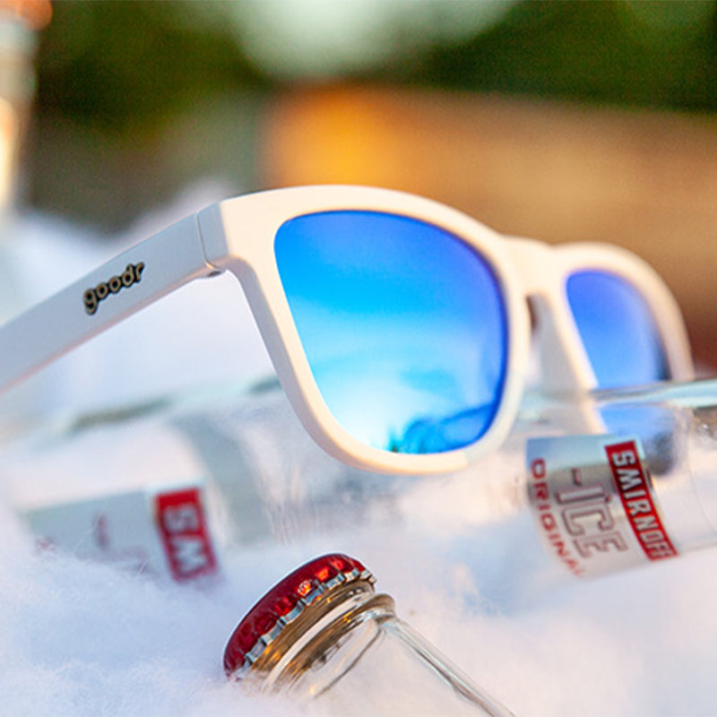 Vista en ángulo de tres cuartos de unas gafas de sol blancas con cristales reflectantes azules sobre un refresco de vodka embotellado en una cubitera.