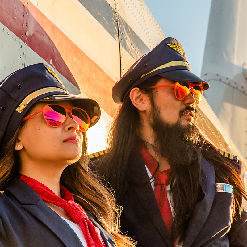 Dos pilotos desaliñados con gafas de sol de aviador con cristales rojos se apoyan en un avión, mirando hacia la puesta de sol.