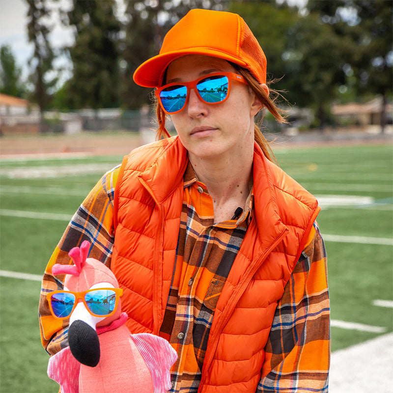 Una mujer vestida de naranja chillón sostiene un flamenco rosa de peluche. Ambas llevan gafas de sol naranjas con cristales azules.