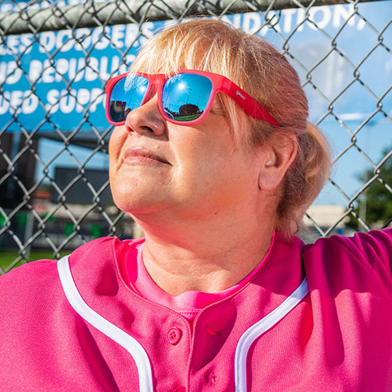 Una mujer con un uniforme de béisbol rosa y unas gafas de sol rosas con cristales azules se apoya en una valla bajo un rayo de sol.