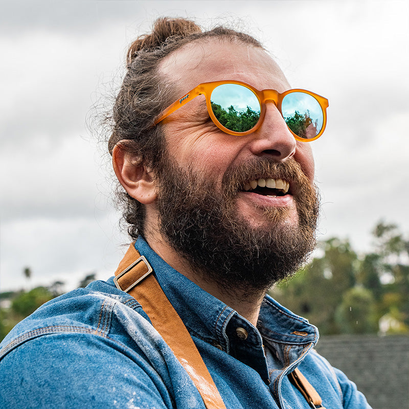 Un hombre barbudo con un moño sonríe con gafas de sol redondas de color naranja con lentes azul claro y un delantal cubierto de harina.