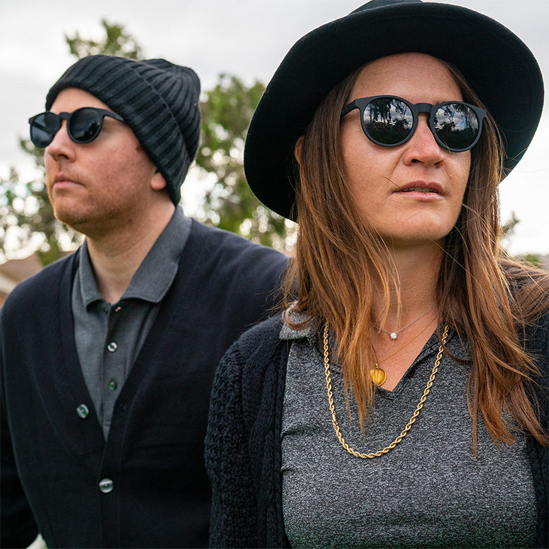 Un hombre y una mujer hipster con elegantes atuendos negros llevan gafas de sol negras redondas con cristales negros antirreflejos.