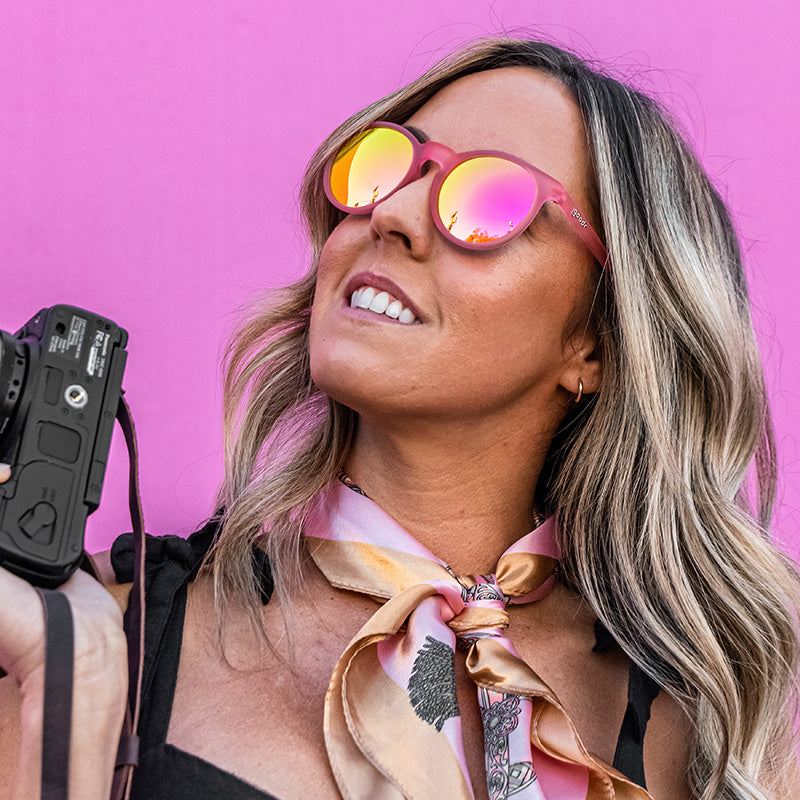 Una elegante mujer con gafas de sol redondas de color rosa y una cámara en la mano mira hacia un lado, con una pared de color rosa intenso a sus espaldas.