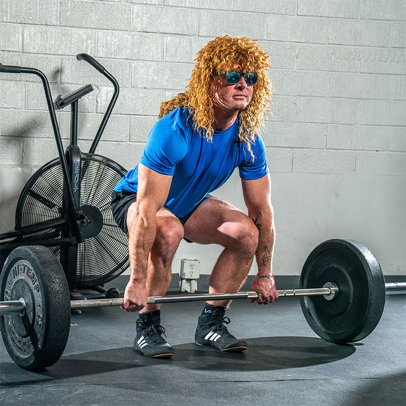 Un hombre con una gran peluca rizada y gafas de sol negras con cristales azules reflectantes levanta una pesada mancuerna en un gimnasio.