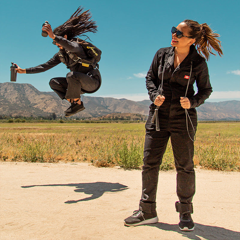 Un paracaidista aterriza en un campo cerca de una mujer con mono negro, ambos llevan gafas de sol negras de aviador.