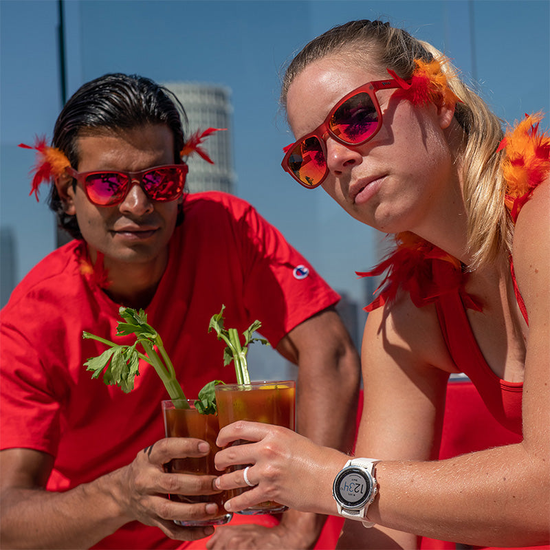 Un hombre y una mujer con plumas naranjas y rojas y gafas de sol rojas con cristales reflectantes rojos sostienen bloody marys en un tejado.