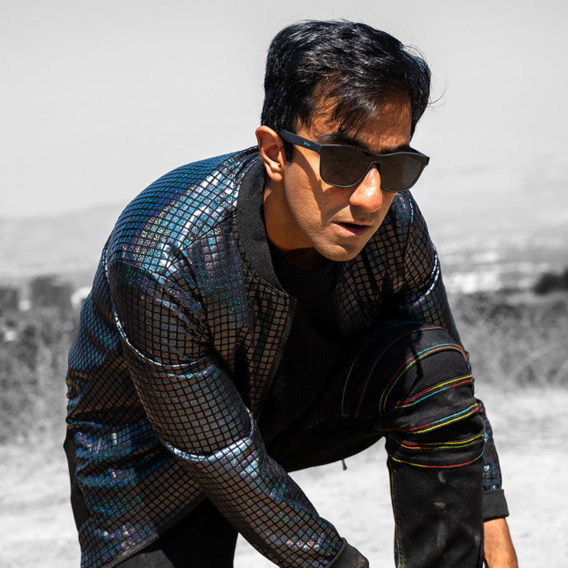 Un hombre con gafas de sol negras planas de una sola lente y una futurista chaqueta reflectante oscura arremete, mirando al frente.