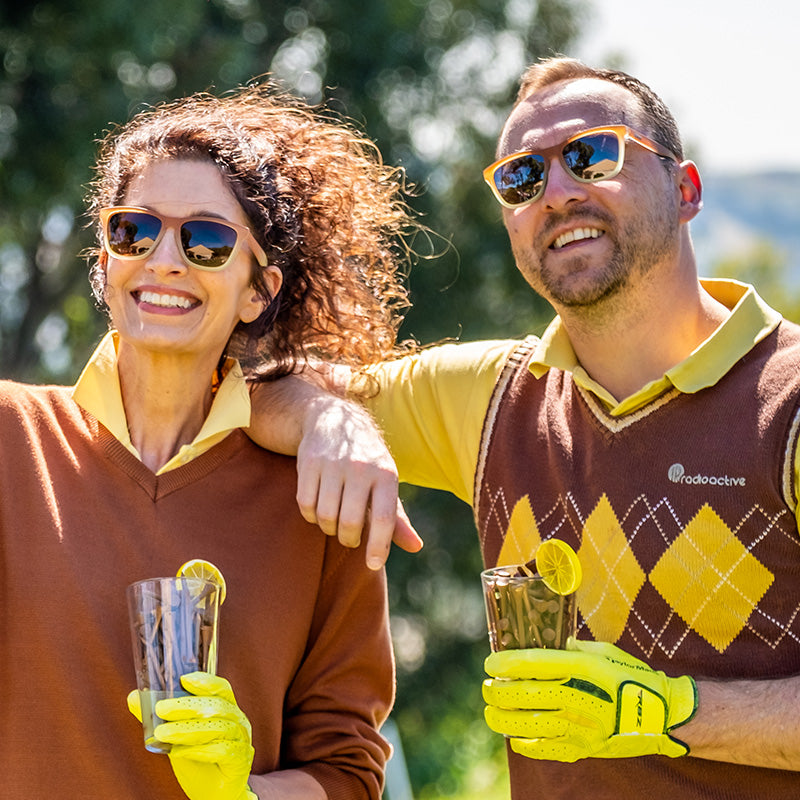 Un hombre y una mujer con gafas de sol degradadas de marrón a blanco con cristales marrones sonríen en un campo de golf sosteniendo un té helado.