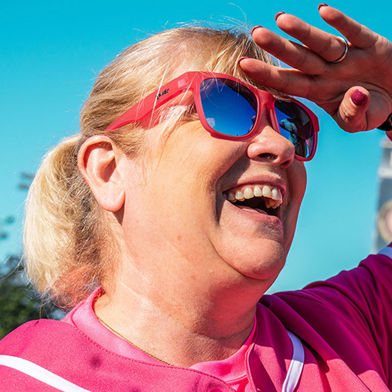 Vista en ángulo de tres cuartos de una señora con uniforme de béisbol rosa y gafas de sol rosas con cristales azules sonriendo en un campo de béisbol.