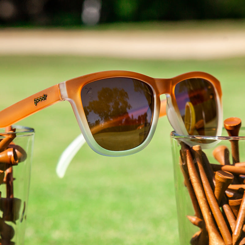Vista en ángulo de tres cuartos de unas gafas de sol con degradado de marrón a blanco colocadas sobre un campo de vasos con tees de golf marrones.