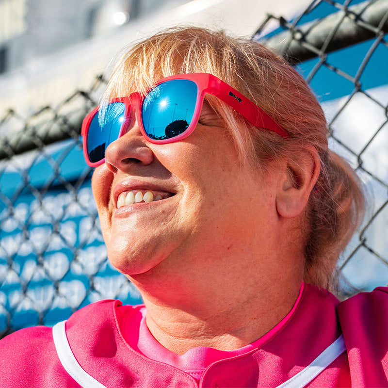 Una mujer con uniforme de béisbol rosa y gafas de sol rosas con cristales reflectantes azules sonríe, apoyada en una valla.