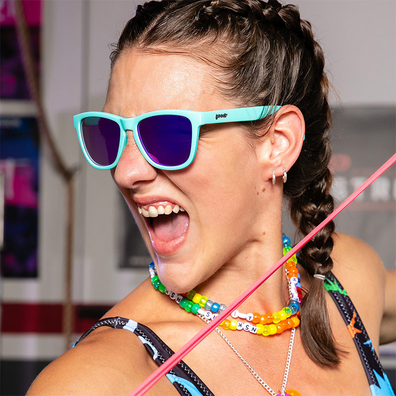Una mujer con collares de plástico arco iris y gafas de sol azul bebé con cristales morados reflectantes gruñe ferozmente en un gimnasio.