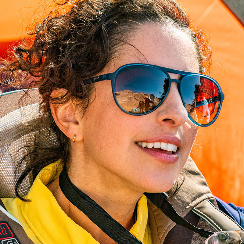 Una mujer con gafas de sol de aviador azul marino con cristales ámbar oscuro sonríe suavemente mirando a lo lejos.
