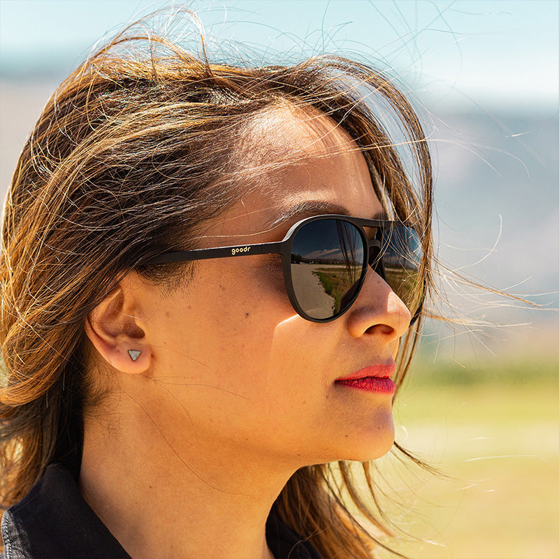 Una mujer con un mono negro y gafas de sol de aviador negras con cristales negros no reflectantes mira al frente con fiereza.