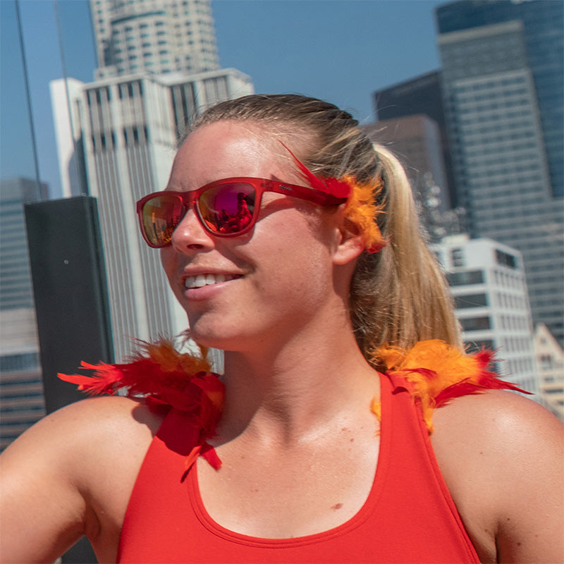 Una mujer lleva gafas de sol rojas con cristales rojos con detalles de plumas rojas y naranjas, el horizonte de una ciudad detrás de ella.
