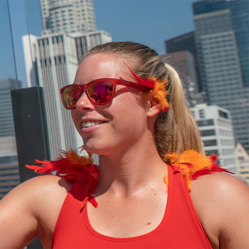Una mujer lleva gafas de sol rojas con cristales rojos con detalles de plumas rojas y naranjas, el horizonte de una ciudad detrás de ella.