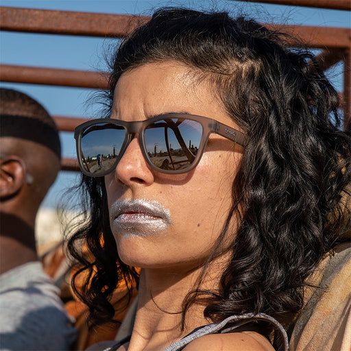 Una mujer feroz con gafas de sol grises con cristales grises y pintura plateada sobre la boca conduce un oxidado coche postapocalíptico.