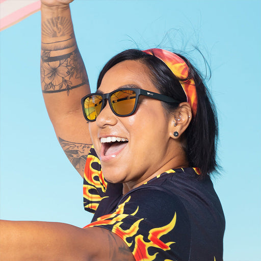 Una mujer con gafas de sol negras con cristales reflectantes ámbar que lleva una camisa con estampado de llamas y una cinta en la cabeza se ríe.